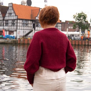 May include: A deep burgundy long-sleeved jumper with a textured knit finish, worn over a light cream-coloured top and paired with off-white trousers. The jumper features a rounded neckline and a relaxed fit, providing a solid colour contrast.
