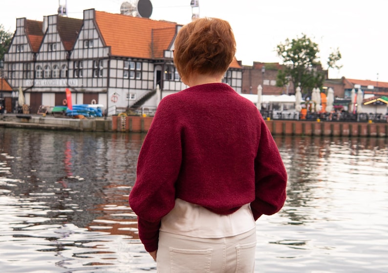 May include: A deep burgundy long-sleeved jumper with a textured knit finish, worn over a light cream-coloured top and paired with off-white trousers. The jumper features a rounded neckline and a relaxed fit, providing a solid colour contrast.