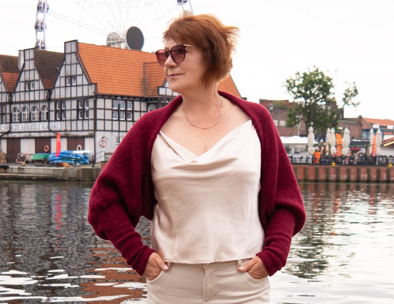 May include: A deep burgundy knit shrug with long sleeves worn over a cream-coloured satin camisole with a cowl neckline. The shrug features a textured knit fabric and is paired with light-coloured trousers, creating a layered look with contrasting textures.