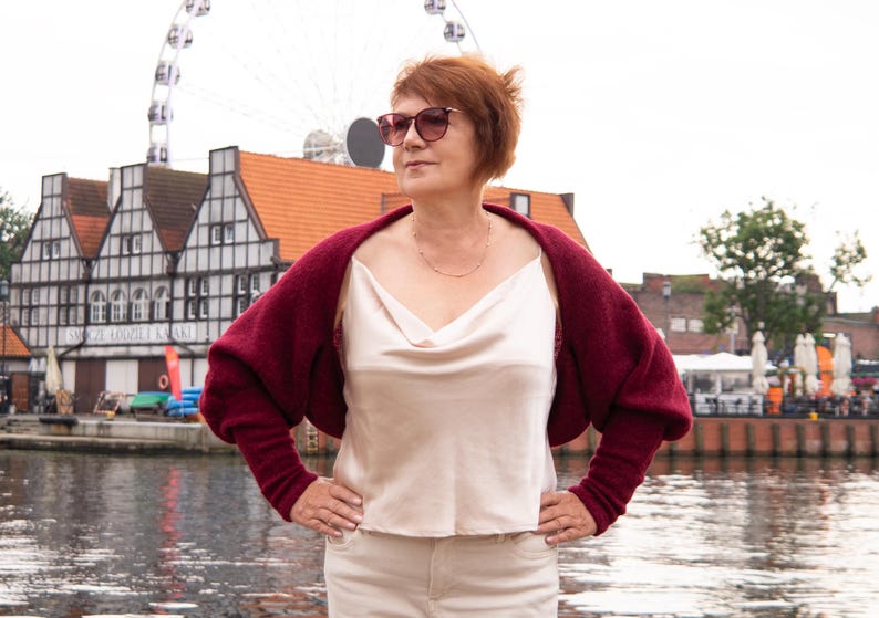 May include: A deep burgundy knit shrug with long sleeves worn over a cream-coloured sleeveless top with a cowl neckline. The shrug features a soft, textured knit fabric and a cropped silhouette that sits across the shoulders and upper back.