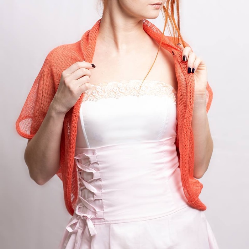 May include: A bright coral orange knit shrug with an open front and short sleeves, worn over a pale pink strapless bodice featuring a lace-trimmed neckline and a corset-style side lacing detail with matching ribbon ties at the waist.