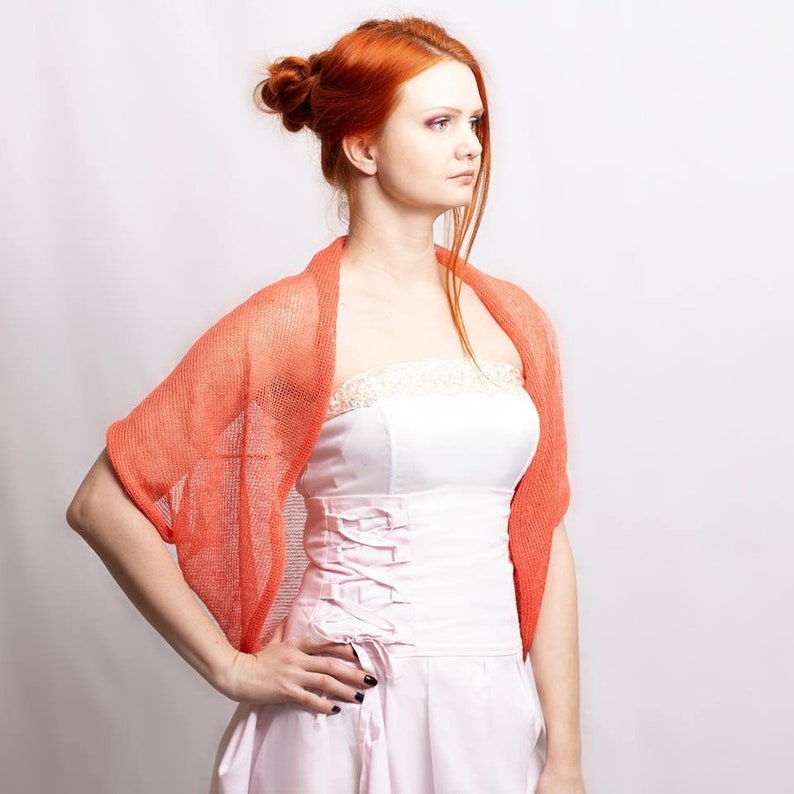 May include: A coral-colored, open-knit shrug with a lightweight, semi-sheer texture worn over a white strapless dress featuring a lace-trimmed neckline and a corset-style bodice with side lacing details and a pleated skirt.