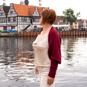 May include: A deep burgundy knit shrug with long sleeves worn over a cream-coloured sleeveless top with a cowl neckline and light-coloured trousers. The shrug features a textured knit fabric and covers the shoulders and arms while leaving the torso exposed.