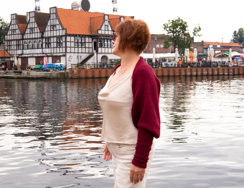 May include: A deep burgundy knit shrug with long sleeves worn over a cream-coloured sleeveless top with a cowl neckline and light-coloured trousers. The shrug features a textured knit fabric and covers the shoulders and arms while leaving the torso exposed.