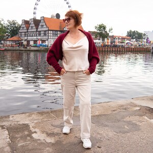 May include: A burgundy knit shrug worn over a cream-coloured satin camisole with a cowl neckline, paired with light beige trousers and white trainers. The outfit includes dark-tinted sunglasses and a thin gold chain necklace.