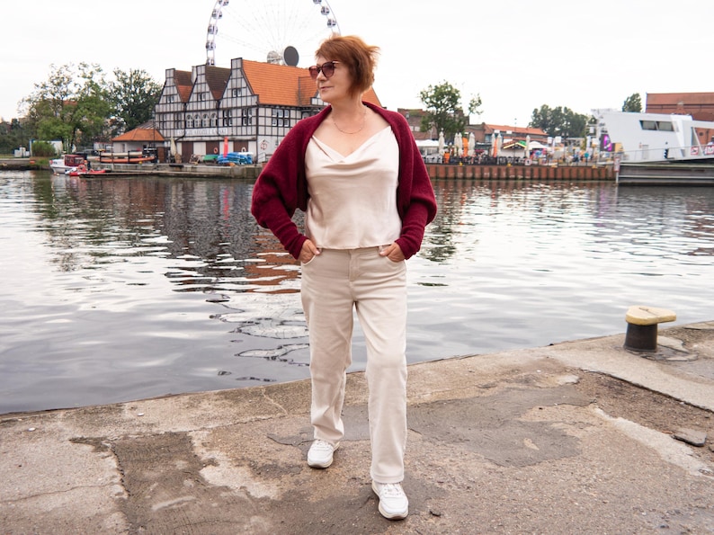 May include: A burgundy knit shrug worn over a cream-coloured satin camisole with a cowl neckline, paired with light beige trousers and white trainers. The outfit includes dark-tinted sunglasses and a thin gold chain necklace.