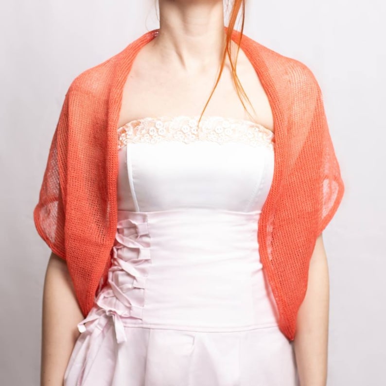 May include: A bright coral orange knit shrug with a loose, open-weave texture worn over a pale pink strapless dress featuring a lace-trimmed neckline and a corset-style bodice with side ribbon lacing details and a pleated skirt section.