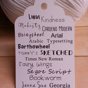 May include: A white tag with a list of fonts written in black ink. The fonts listed are Luna, Kindness, Modesty, Gardeno Modern, Bringshoot, Arial, Arabic Typesetting, Barthowheel, Tommy's, Sketched, Times New Roman, Fairy Wings, Segoe Script, Bookworm, Jenna Sue, and Georgia.