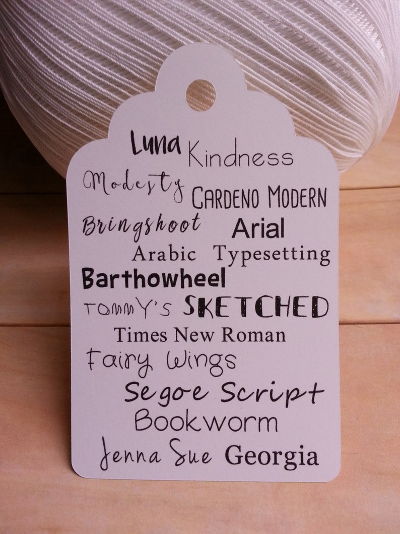 May include: A white tag with a list of fonts written in black ink. The fonts listed are Luna, Kindness, Modesty, Gardeno Modern, Bringshoot, Arial, Arabic Typesetting, Barthowheel, Tommy's, Sketched, Times New Roman, Fairy Wings, Segoe Script, Bookworm, Jenna Sue, and Georgia.