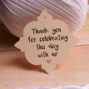 May include: A beige tag with a decorative border and a hole at the top. The tag has the text "Thank you for celebrating this day with us!" written in black cursive with a small pink heart below the text.