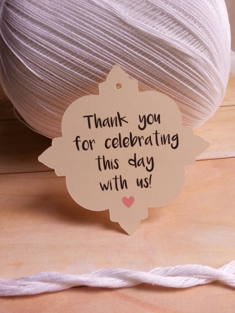 May include: A beige tag with a decorative border and a hole at the top. The tag has the text "Thank you for celebrating this day with us!" written in black cursive with a small pink heart below the text.