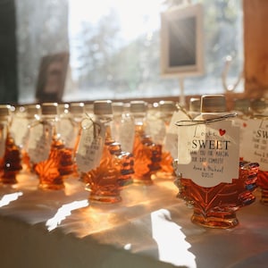 May include: A row of small maple syrup bottles with leaf-shaped glass containers. Each bottle has a tag tied around the neck with the text "Love is Sweet" and a small red heart. The tags thank guests for making the wedding day complete.