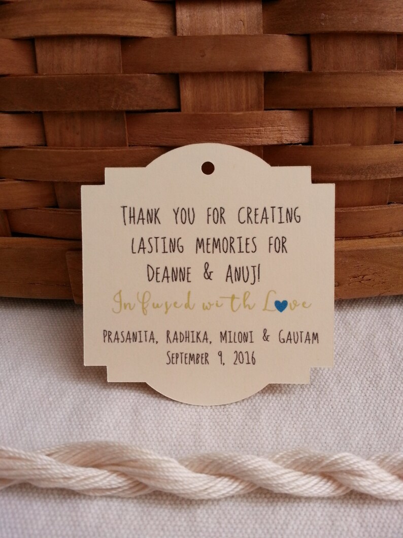 May include: A cream-colored thank you tag with a rounded top and a blue heart. The tag reads "Thank you for creating lasting memories for Deanne & Anuj! In fused with love. Prasanita, Radhika, Miloni & Gautam. September 9, 2016."