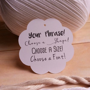 May include: White cloud-shaped tag with black text that reads "Your Phrase! Choose a Shape! CHOOSE A SIZE! Choose a Font!" The tag is sitting on a wooden surface with white, cream, and beige yarn.