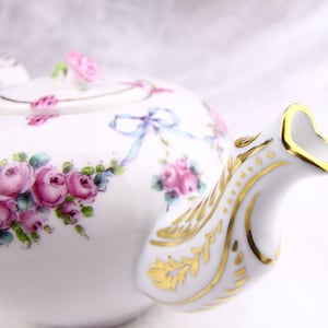 Peut inclure: Une théière en porcelaine blanche avec un motif floral doré et un ruban bleu. La théière a un bec en forme de cœur.