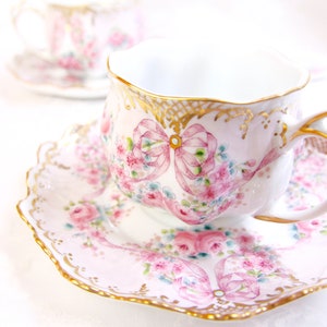 May include: A delicate teacup and saucer set with a pink and white floral design and gold trim. The cup and saucer feature a pink ribbon design with a floral pattern.