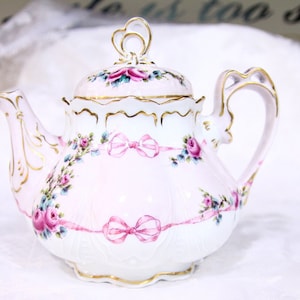 Peut inclure: Une théière en porcelaine blanche avec un motif floral rose et des accents dorés. La théière a un couvercle avec une poignée en forme de cœur et un bec verseur avec une courbe délicate. La théière est décorée de roses roses, de feuilles vertes et de rubans roses.