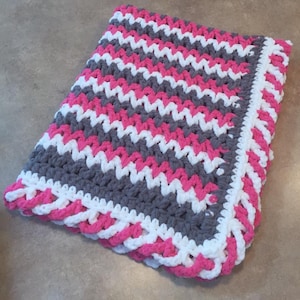 May include: A crocheted baby blanket in gray, white, and pink with a chevron pattern. The blanket has a pink and white border.