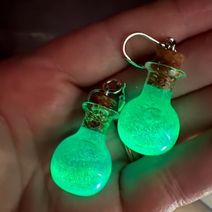 May include: A pair of glowing green potion bottle earrings. Each glass bottle has a cork stopper and a silver-colored ear wire. The earrings are filled with a glowing green liquid, creating a magical effect.