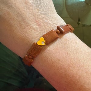 May include: A brown bracelet with a gold heart charm and a small clear stone in the center. The bracelet is made of small, rectangular brown stones.