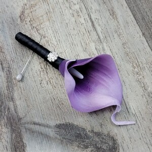 May include: A boutonniere featuring a single purple calla lily flower with a white spadix. The stem is wrapped in black ribbon and accented with a small, circular, silver-toned rhinestone embellishment. A silver pin with a pearl-like bead is attached.
