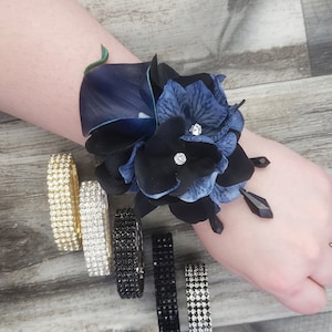 May include: A wrist corsage featuring dark blue and black artificial flowers with small clear rhinestones and black teardrop beads, accompanied by five separate elastic bracelets in gold, silver, black, and clear rhinestone finishes.