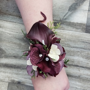 May include: A wrist corsage featuring a deep burgundy calla lily, dark purple hydrangea petals, and small cream-colored flowers. The arrangement is accented with sprigs of green cedar foliage and small clear rhinestones centered on the petals.