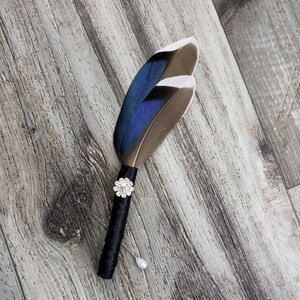 May include: A handcrafted boutonniere featuring two feathers with blue and brown hues, accented with a sparkling rhinestone embellishment and a black satin-wrapped stem. Includes a pearl-headed pin for secure attachment. A unique accessory for weddings or special events.