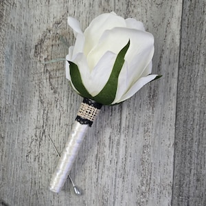 May include: A white rose boutonniere with green leaves and a black and beige decorative band. The stem is wrapped in white satin ribbon and secured with a silver pin. The flower is set against a gray wooden background.