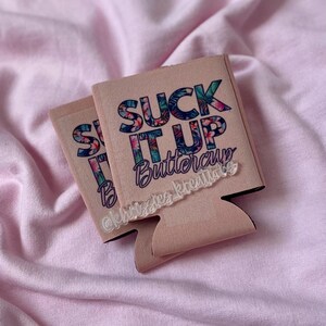 May include: Two light pink can coolers with the text "SUCK IT UP Buttercup" in a floral design. The text is in blue, pink, and green. The bottom of the cooler has a black trim. The background is a light pink fabric.