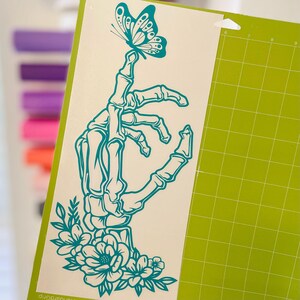 May include: Teal blue skeleton hand with floral details and a butterfly perched on its finger. The design is on a white background.