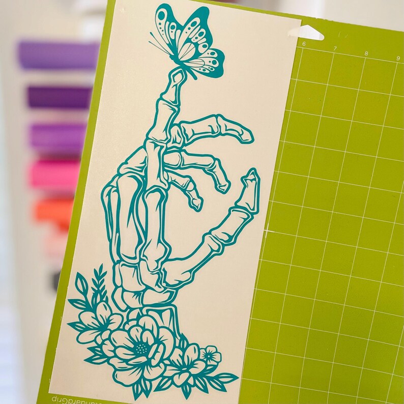May include: Teal blue skeleton hand with floral details and a butterfly perched on its finger. The design is on a white background.