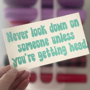 May include: A rectangular sticker with the text "Never look down on someone unless you're getting head" in light blue, retro-style font. The sticker is held in a hand, with a blurred background of shelves and objects.