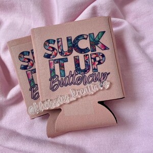 May include: Two pink can coolers with the text "SUCK IT UP Buttercup" in a floral design. The coolers have a black bottom and a clear acrylic tag that says "@khizzies kreatiions".