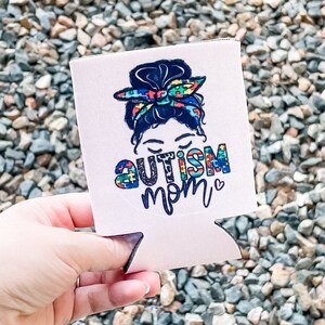 May include: White can cooler featuring a colourful graphic of a woman with a bun in her hair. The woman is wearing a headband with jigsaw pieces. The text "AUTISM MOM" is printed below the woman's head.