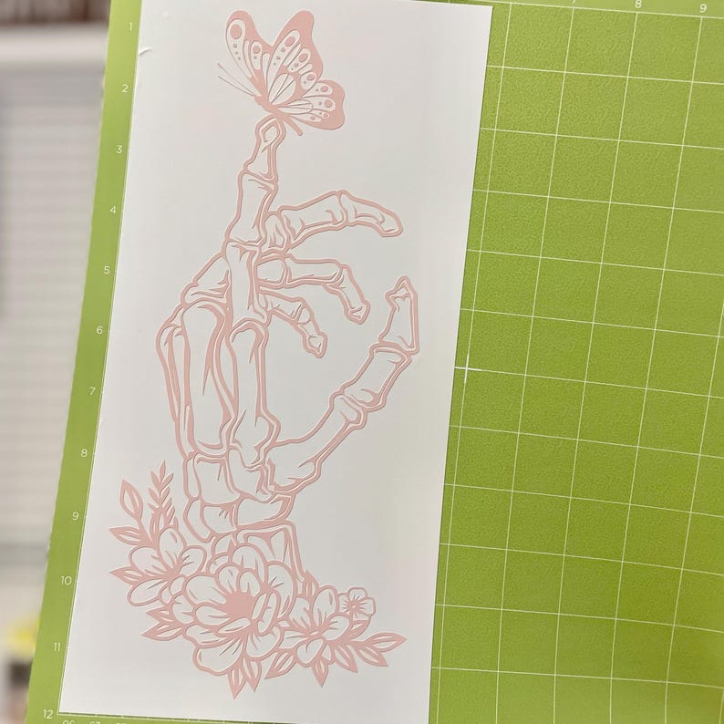 May include: A pink skeleton hand with a butterfly perched on its fingertip. The hand is surrounded by pink flowers and leaves.