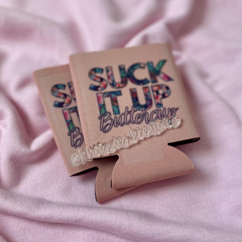 May include: Two blush-colored drink koozies with the text "SUCK IT UP Buttercup" in a floral design. The koozies have a black trim and a cut-out for the bottom of the can. The text "@khizzieskreations" is also visible.
