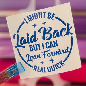 May include: A white sticker with blue text that reads "I MIGHT BE Laid Back BUT I CAN Lean Forward REAL QUICK" in a circular design. The sticker is held by a blue and patterned clip.
