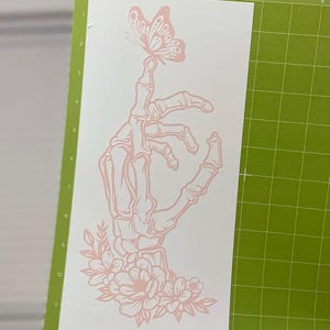May include: A pink skeletal hand with a butterfly perched on its fingertip. The hand is surrounded by pink flowers and leaves.