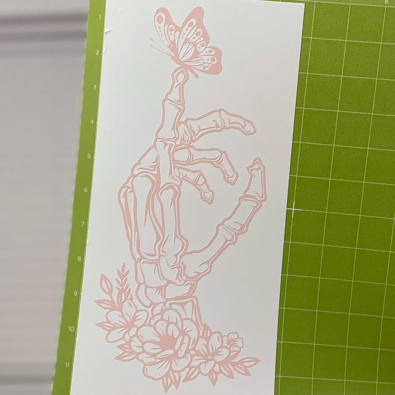 May include: A pink skeletal hand with a butterfly perched on its fingertip. The hand is surrounded by pink flowers and leaves.