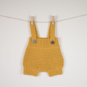 May include: A mustard yellow crocheted baby romper with straps and two silver buttons. The romper is hanging on a clothesline with wooden pegs.