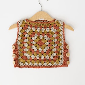 May include: A handmade crochet vest in a granny square pattern, featuring shades of rust, mustard yellow, and cream. The vest has a round neckline and is secured with wooden buttons. It hangs on a wooden hanger against a white background.