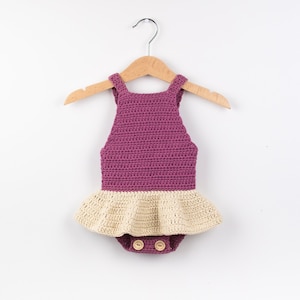 May include: A crocheted baby romper with a pink bodice and a cream-coloured ruffle skirt. The romper has two wooden buttons at the bottom.