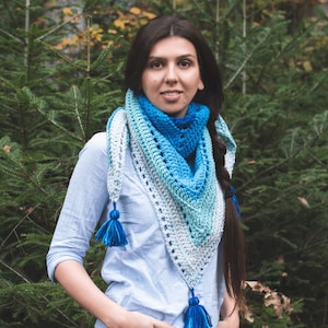 May include: A woman wearing a blue and white crocheted shawl with tassels. The shawl is a gradient of blue and white colours. The woman is standing in a forest.