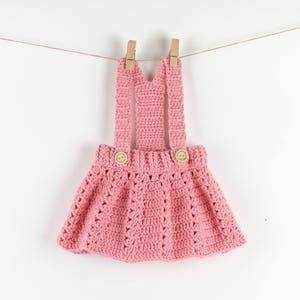 Pode incluir: Um vestido de beb&eacute; de crochet rosa com al&ccedil;as e dois bot&otilde;es de madeira. O vestido est&aacute; pendurado num varal com prendedores de roupa de madeira.