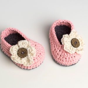 May include: A pair of pink crocheted baby shoes with a white crocheted flower and a brown button on each shoe.