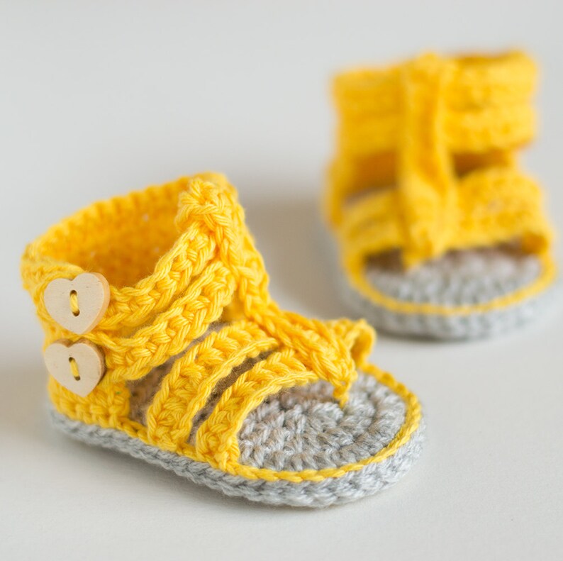 May include: A pair of yellow crocheted baby sandals with gray soles and wooden heart-shaped buttons.