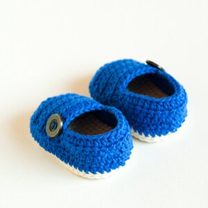 May include: A pair of blue crocheted baby shoes with white trim and a brown button on each shoe.