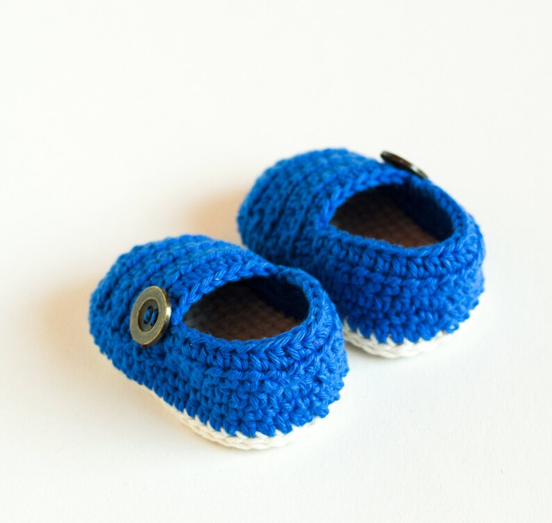 May include: A pair of blue crocheted baby shoes with white trim and a brown button on each shoe.