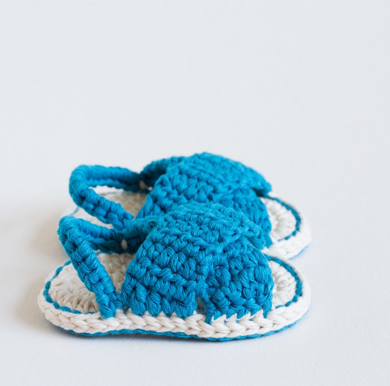May include: A pair of crocheted baby sandals in blue and white. The sandals have a simple design with a strap across the top of the foot.